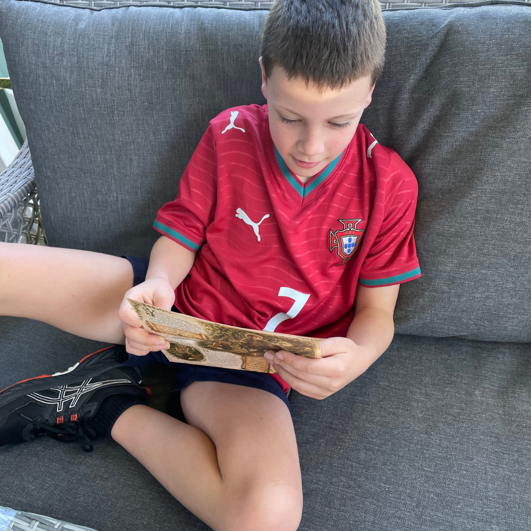 A boy sitting on a gray outdoor sofa wearing a red Portugal Puma soccer jersey and holding a piece of paper.