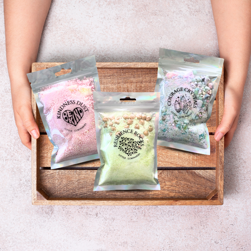 Three packages labeled 'Kindness Dust', 'Resilience Box', and 'Courage Crystal' held by a person over a wooden tray.