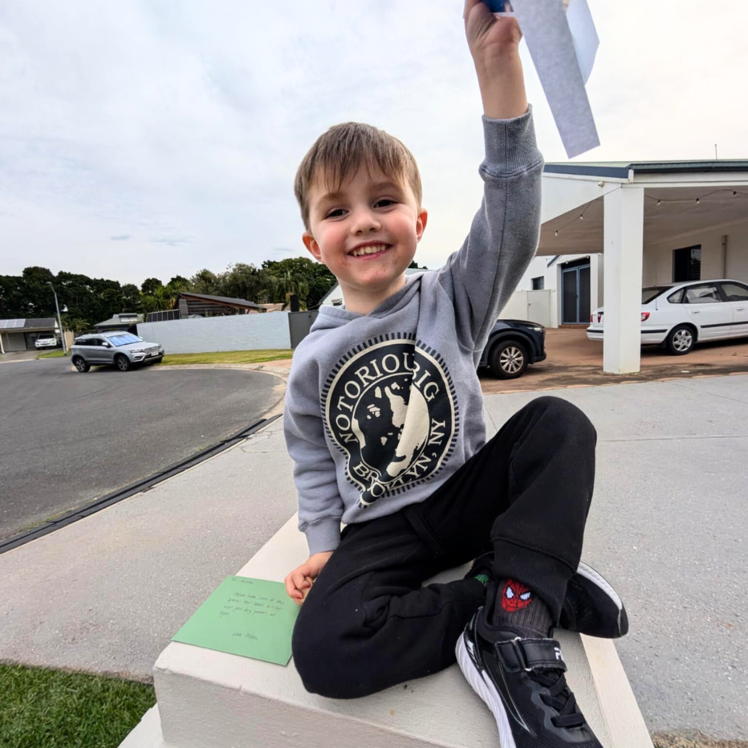 Child sitting on a bench outdoors, holding up a piece of paper with a building and street in the background.