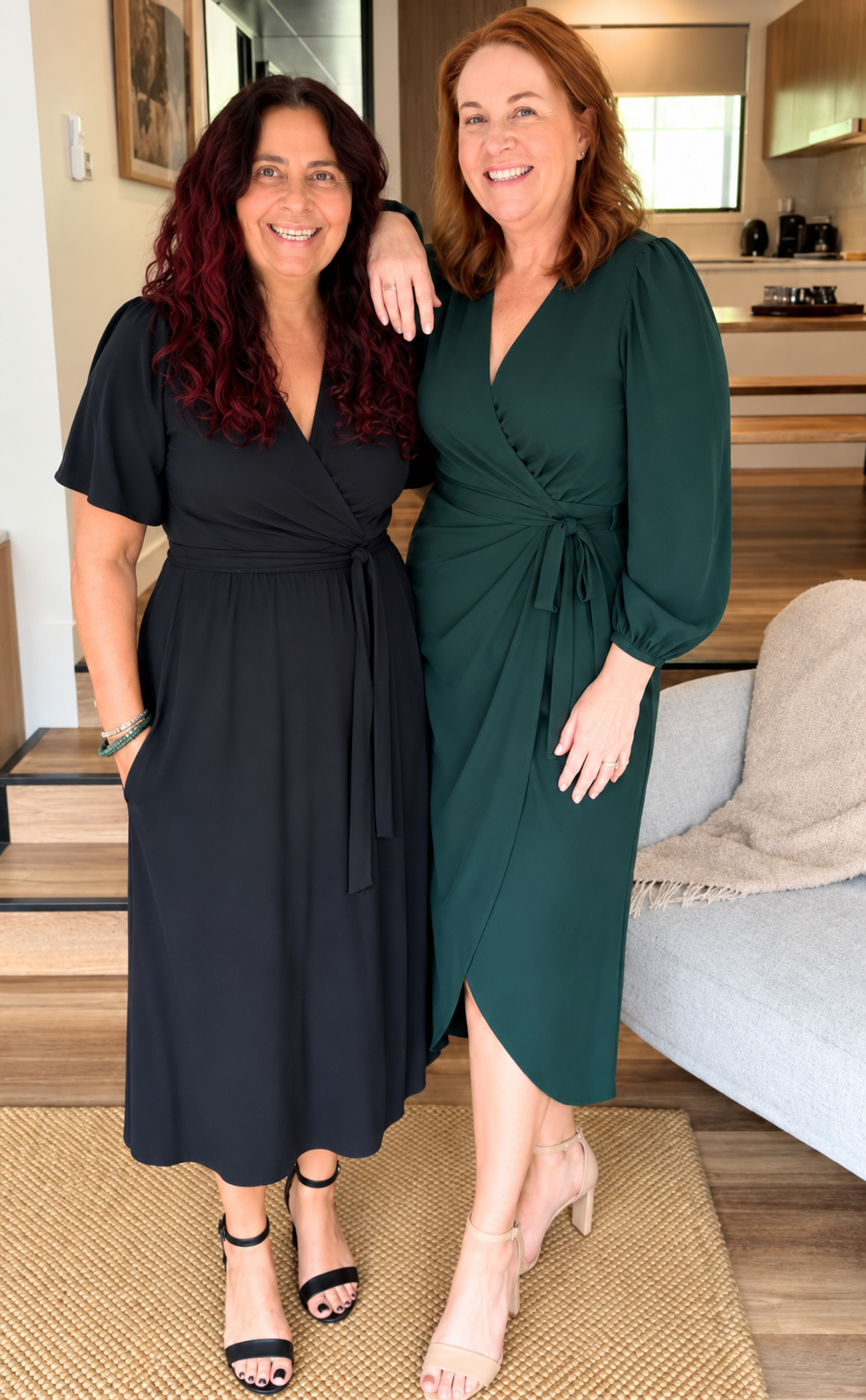 Two women posing indoors wearing midi wrap dresses, one in black and the other in dark green, standing on a woven rug in a modern living space.