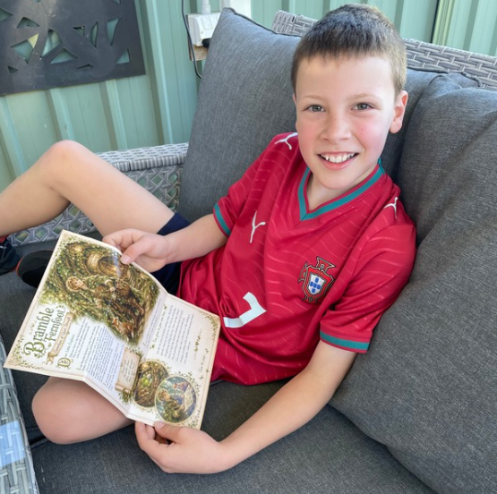 A young boy smiling while sitting on a gray outdoor patio sofa, holding an open book. He is wearing a red Portugal soccer jersey with green trim.