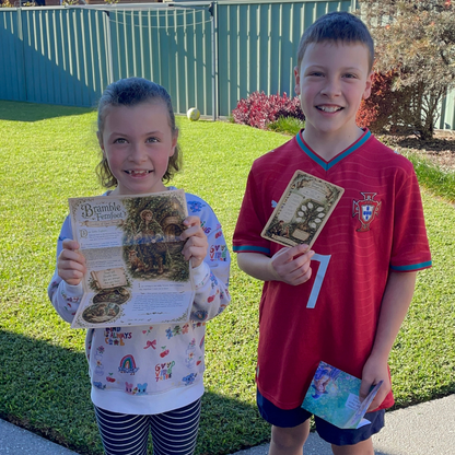 Two children standing outdoors on a lawn in front of a green fence, each holding up a printed book or booklet. The girl wears a white long-sleeve shirt with colorful cartoon patterns and striped pants, and the boy wears a red soccer jersey with a Portugal crest.