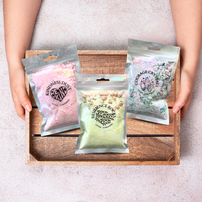 Three packages labeled 'Kindness Dust', 'Resilience Box', and 'Courage Crystal' held by a person over a wooden tray.